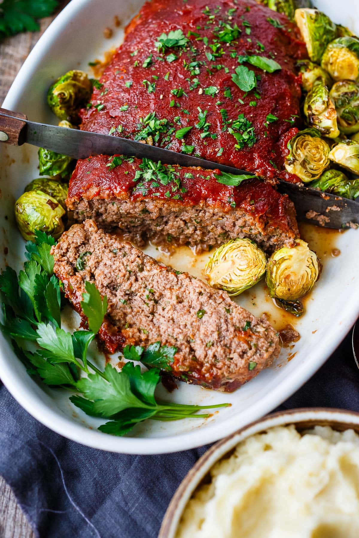 slicing into homemade meatloaf in baking dish surrounded by brussels sprouts and garnished with fresh parsley.