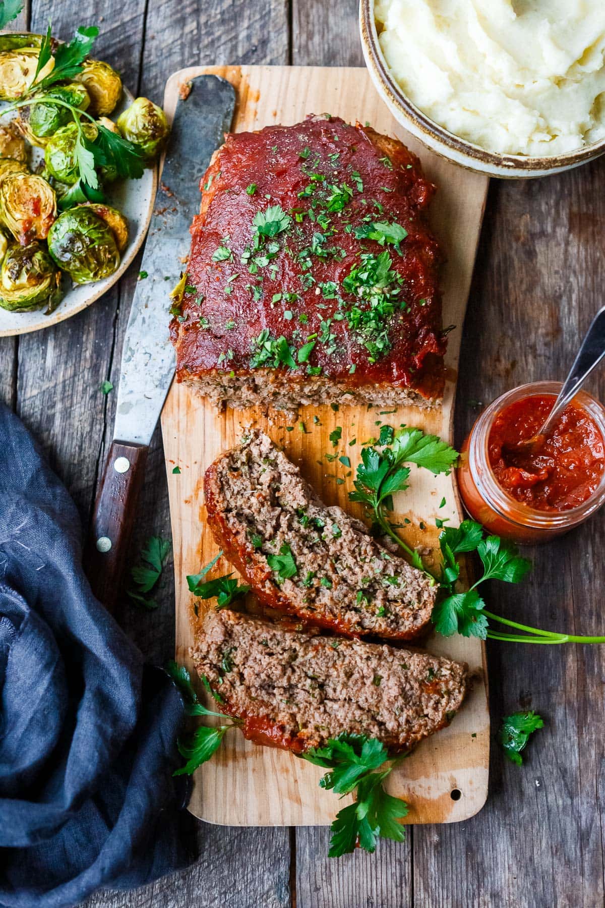 homemade meatloaf on wood board with slices, garnished with fresh parsley, next to mashed potatoes, brussels sprouts, and jar of ketchup. 