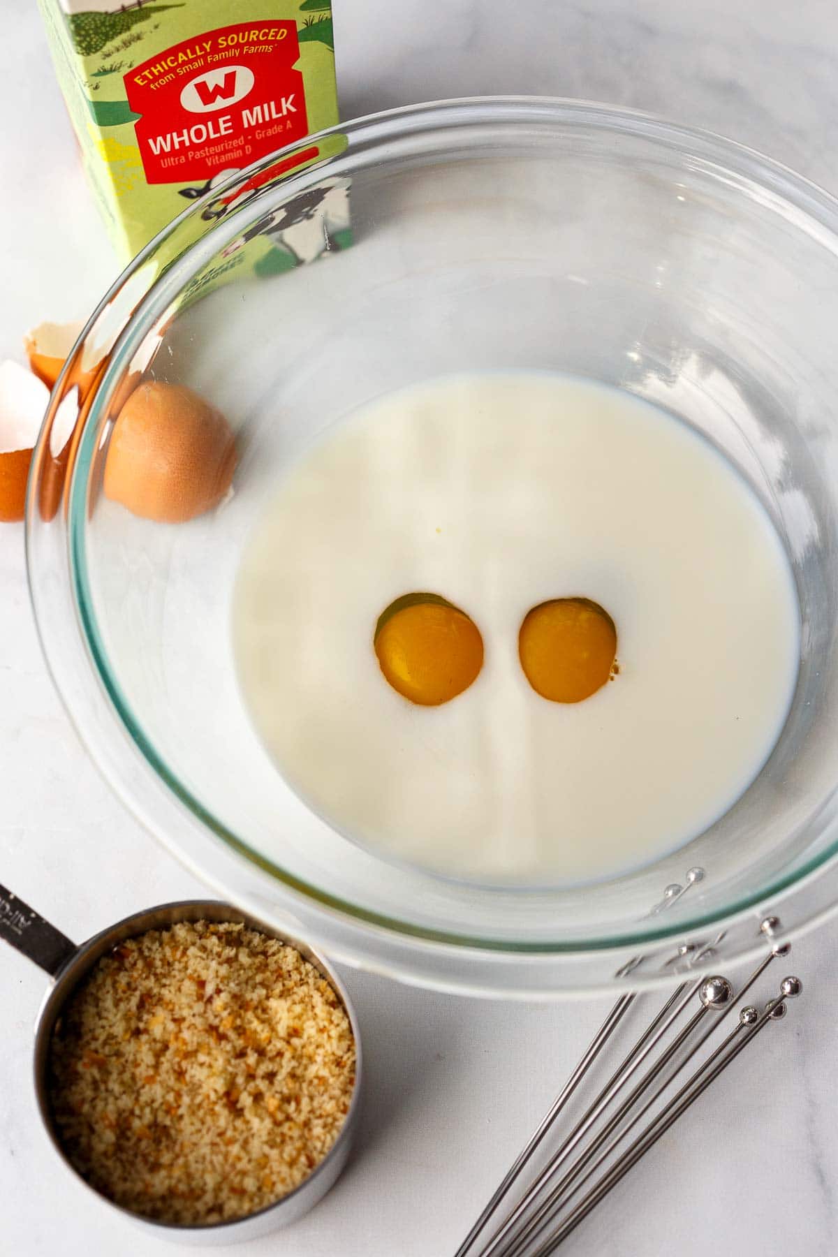 eggs and milk in glass mixing bowl next to cup of breadcrumbs. 