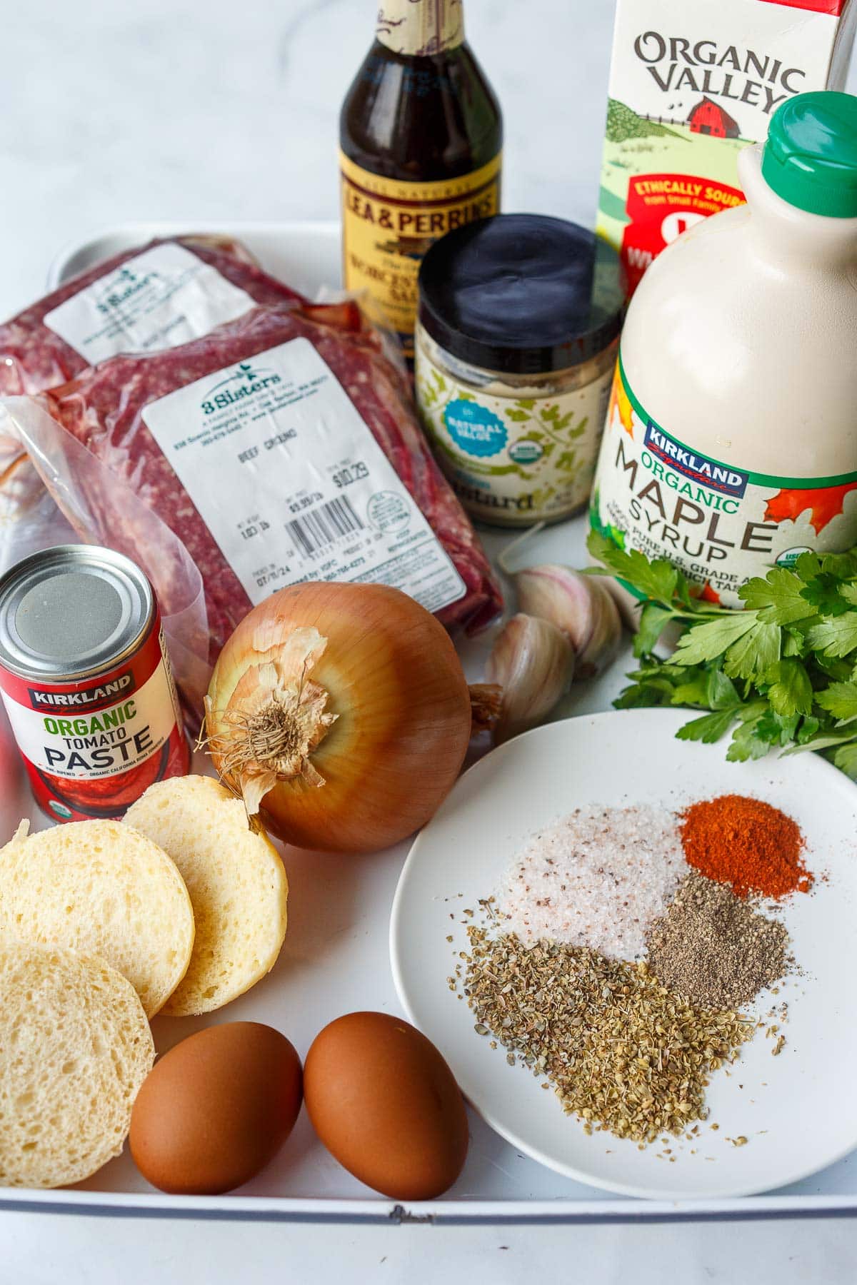 ingredients for homemade meatloaf laid out - ground beef, worcestershire, milk, mustard, maple syrup, parsley, garlic, onion, tomato paste, bread slices, eggs, spices. 