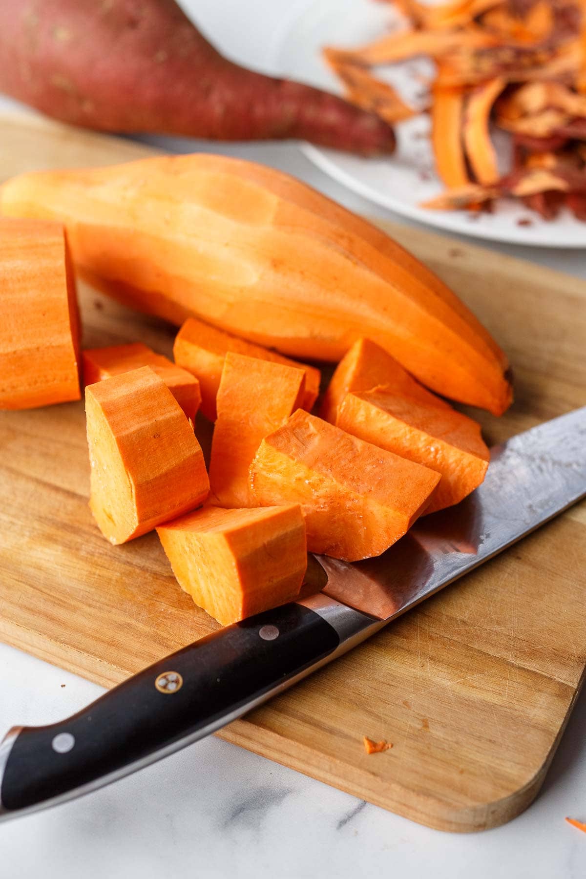 knife on cutting board with chopped sweet potatoes and peeled sweet potato.