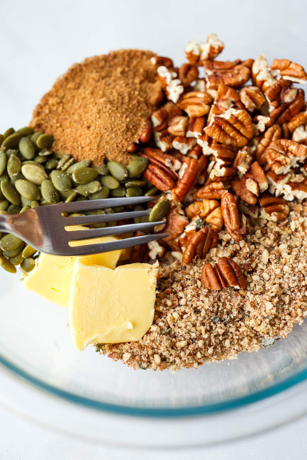 glass mixing bowl with pecan crumble mixture, chopped pecans, butter, pepitas, and spices.