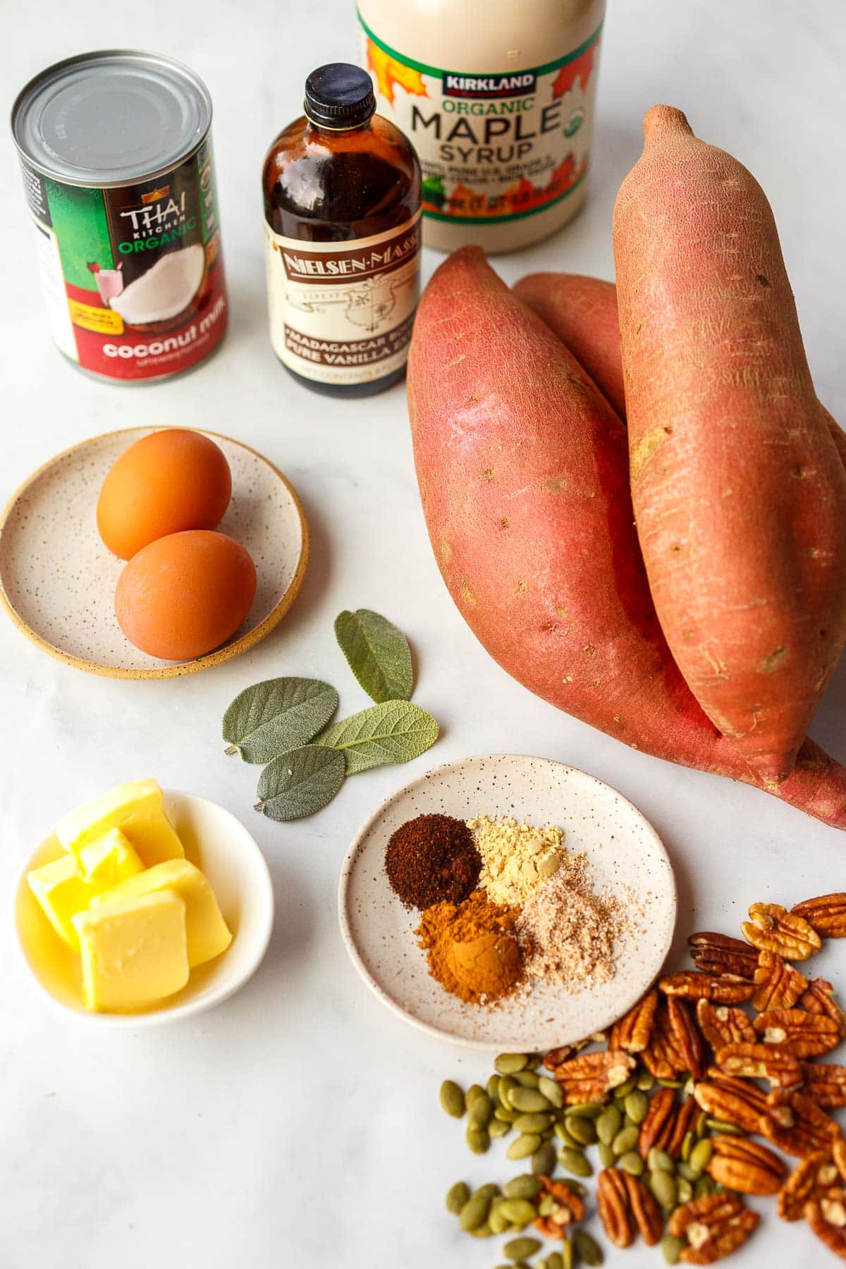 ingredients for sweet potato casserole - maple syrup, vanilla, coconut milk, eggs, sweet potatoes, sage leaves, butter, spices, pecans, pepitas.