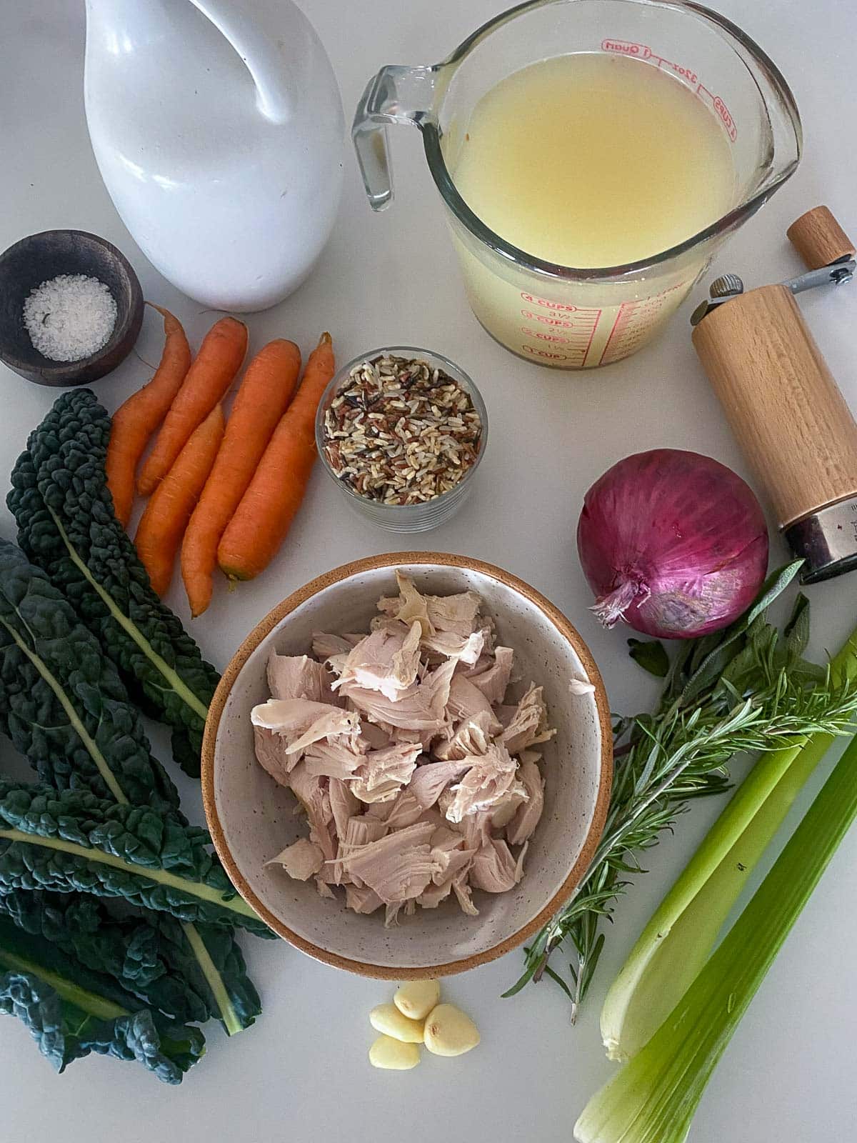 ingredients for turkey rice soup- broth, salt, pepper shaker, wild rice, red onion, carrots, lacinato, bowl of leftover turkey, fresh herbs, celery, garlic.