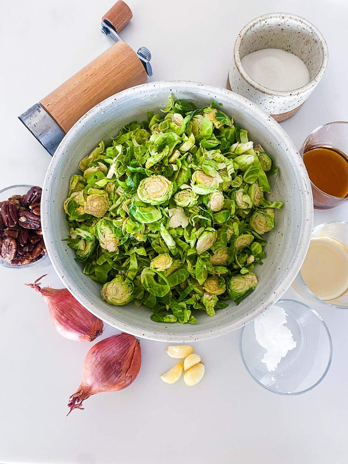 ingredients for sauteed Brussels sprouts laid out- bowl of shredded Brussels sprouts, pepper shaker, bowl of salt, maple syrup, apple cider vinegar, coconut oil, garlic cloves, shallot, and pecans. 