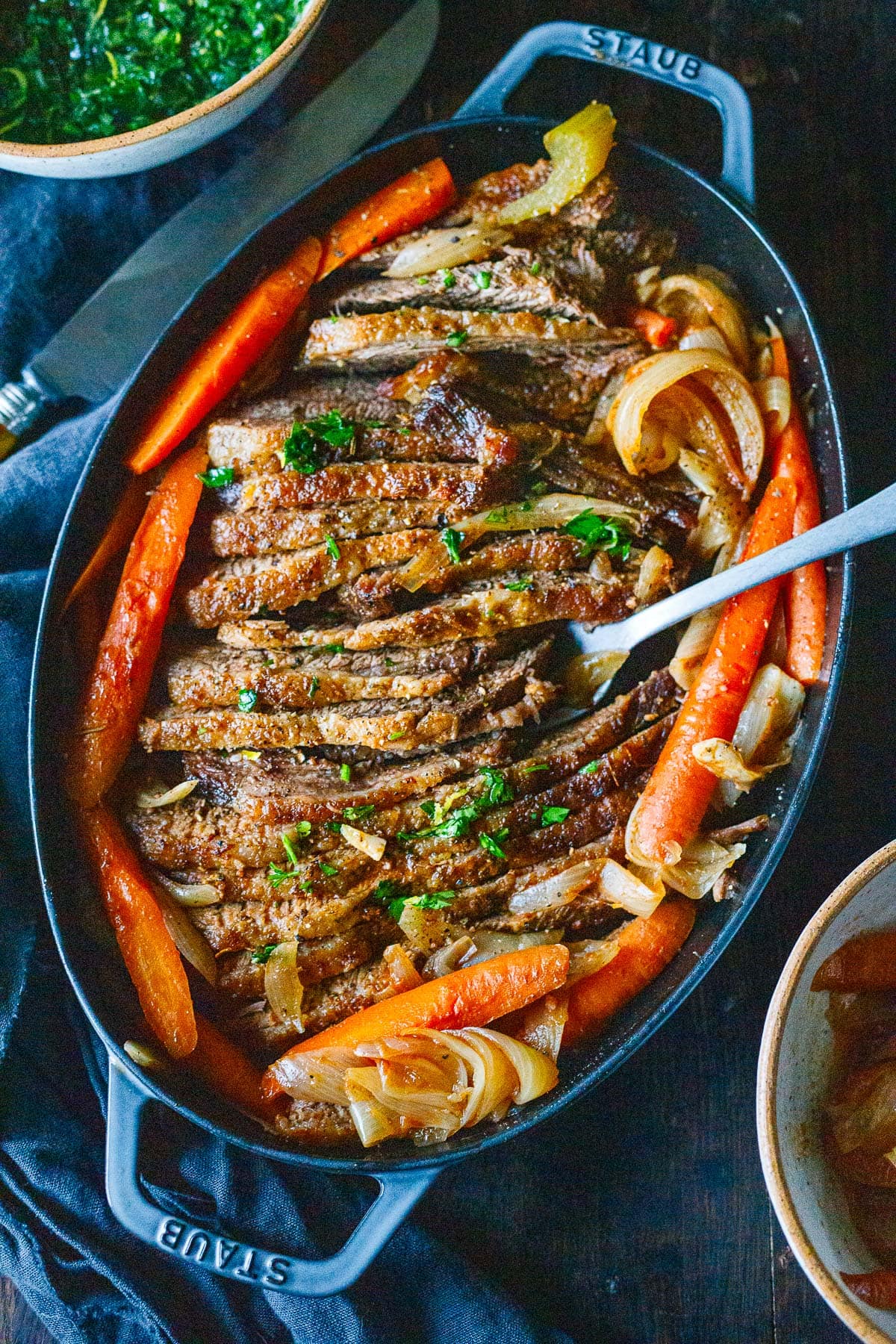 oval baking dish with oven brisket sliced in pan surrounded by onions and carrots. 