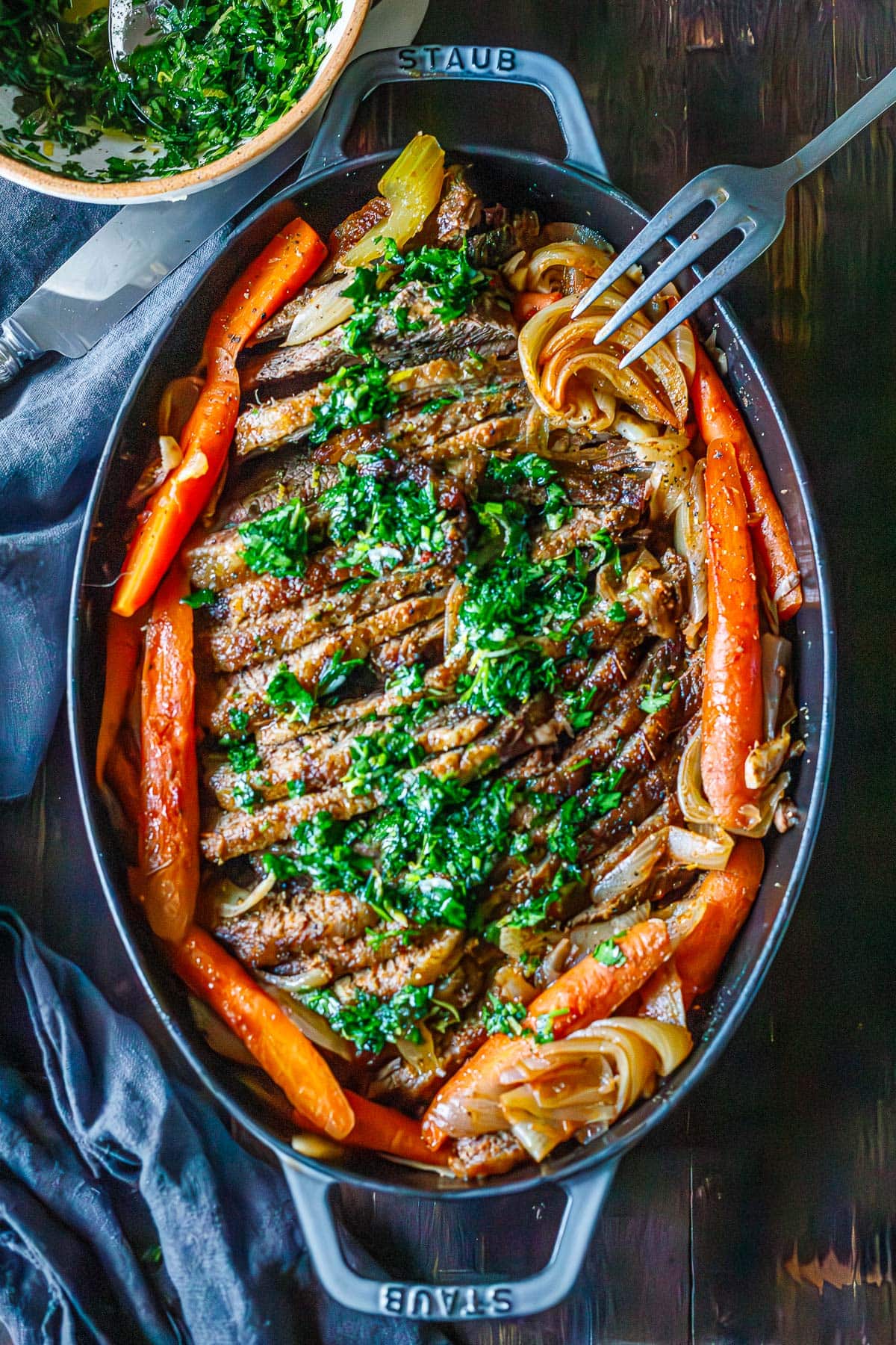 sliced oven brisket in oval dish surrounded by carrots and onions and topped with gremolata. 