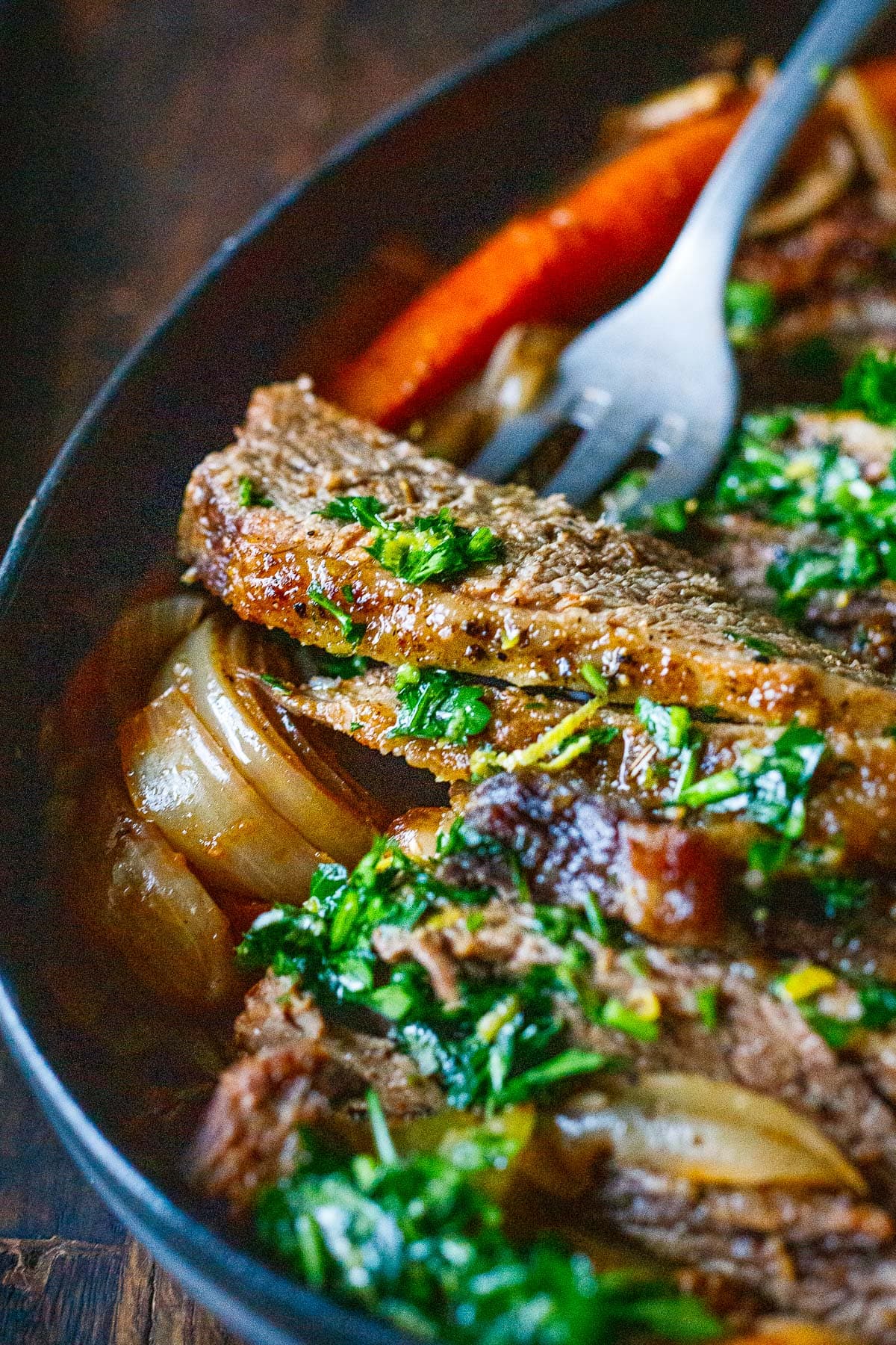 slice of oven brisket with lemon parsley gremolata in baking dish with onions and carrots and fork picking up a piece. 