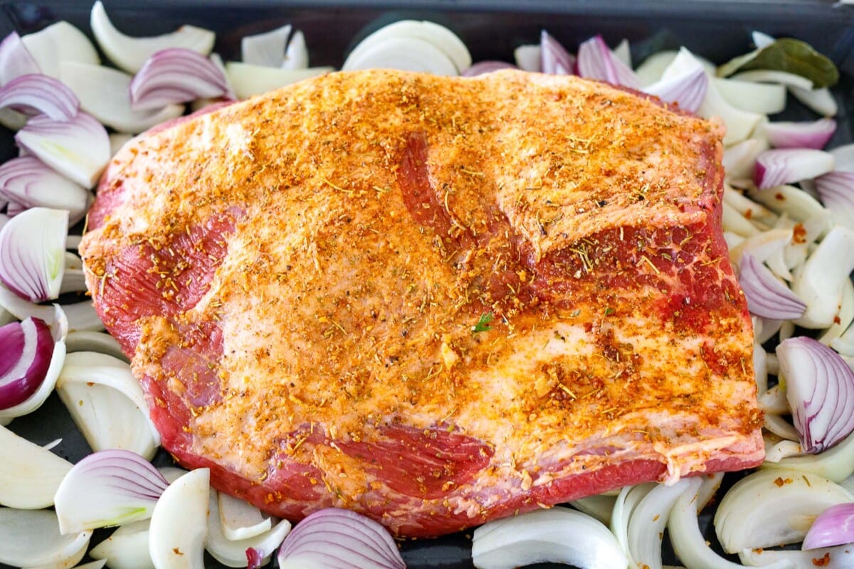 seasoned beef brisket uncooked on baking dish surrounded by onions. 