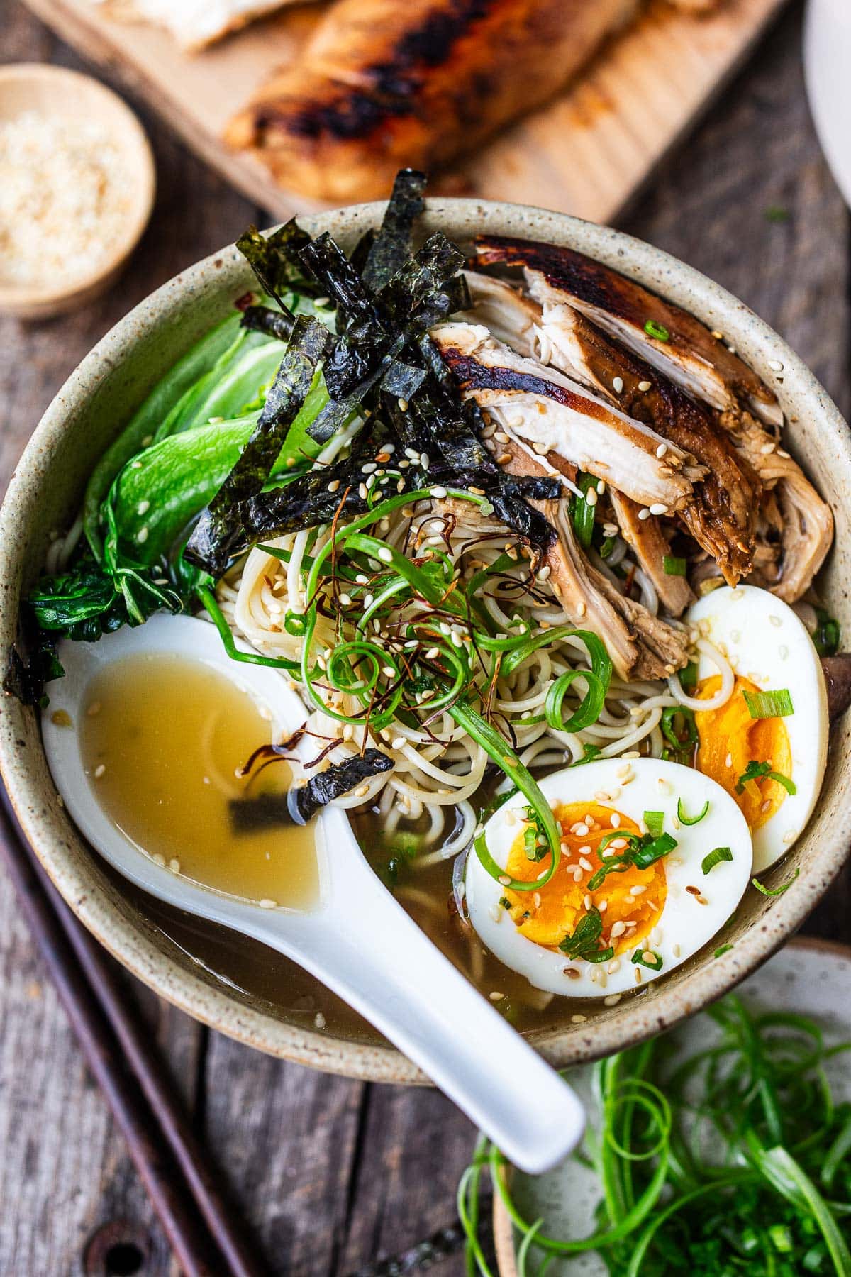 homemade ramen with roasted chicken, boiled eggs, bok choy, nori, scallions, and sesame seeds and spoon resting in broth.