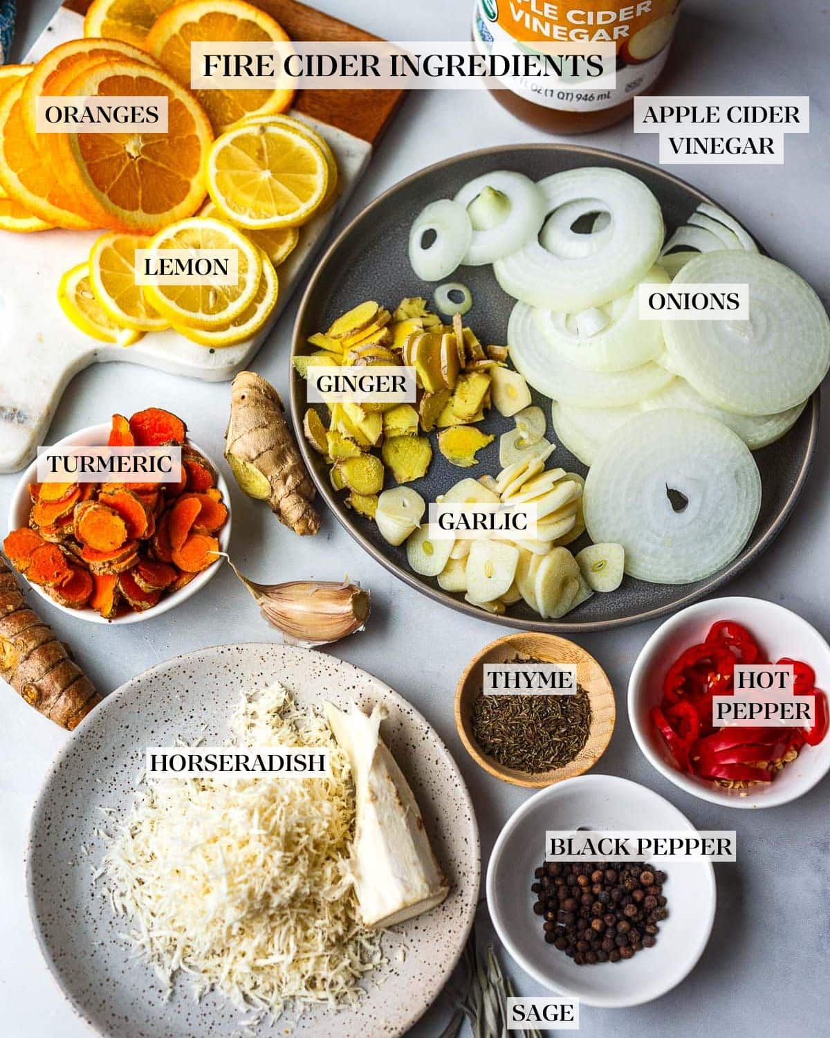 ingredients labeled and spread out for fire cider - apple cider vinegar, sliced citrus, turmeric and ginger root, garlic and onion, hot peppers, black peppercorns, thyme and sage, horseradish grated. 