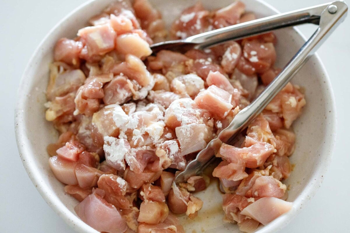 bowl of cubed chicken tossed in cornstarch and soy sauce with tongs.