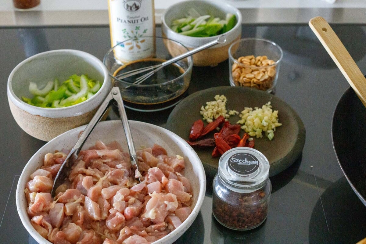 getting ready to cook kung pao chicken - mis en place with bowl of marinated chicken and tongs, szechuan, plate of garlic, ginger, and chiles, jar of peanuts, kung poa sauce, and bowls of celery and scallions next to peanut oil.