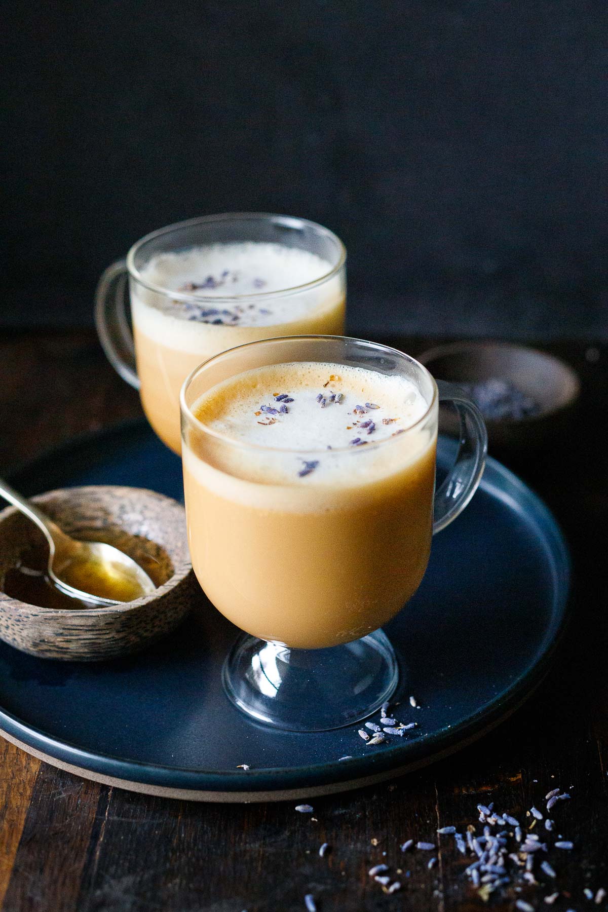 london fog latte in glass mugs with frothy milk and dried lavender garnish on round plate with small bowl of homemade syrup with spoon. 