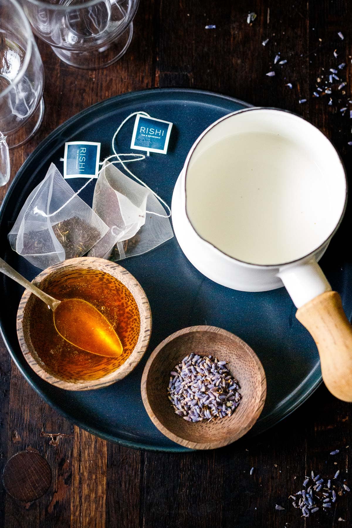 ingredients on round plate for london fog latte - earl grey tea bags, small saucepan with milk, small bowl and spoon with simple syrup, and small bowl with dried lavender.