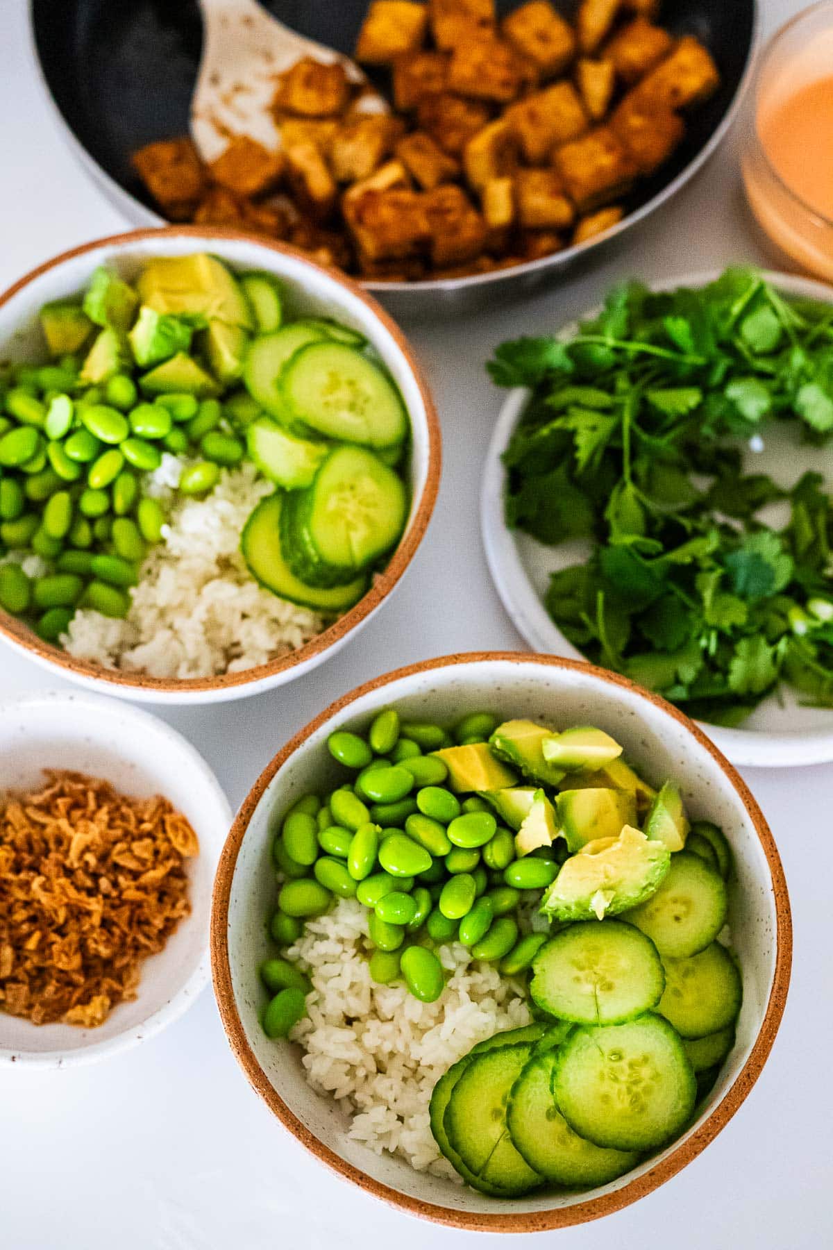 assembling sticky glazed tofu bowls - two bowls of sushi rice, edamame, avocado, and cucumber next to cilantro, crispy onions, and pan-fried tofu.