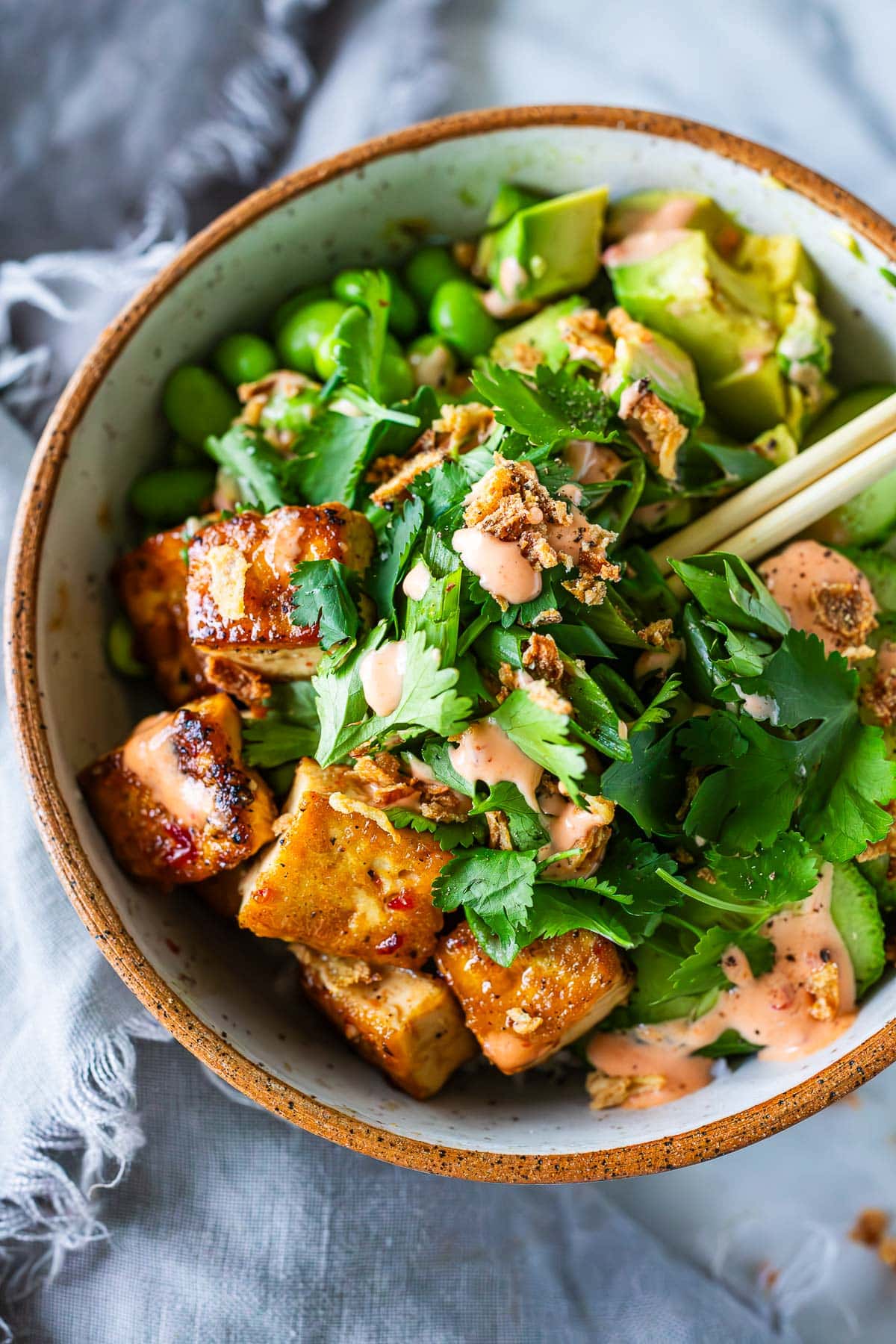 chopsticks in sticky glazed tofu bowl with edamame, avocado, cilantro, and crispy onions, drizzled with yum yum sauce.