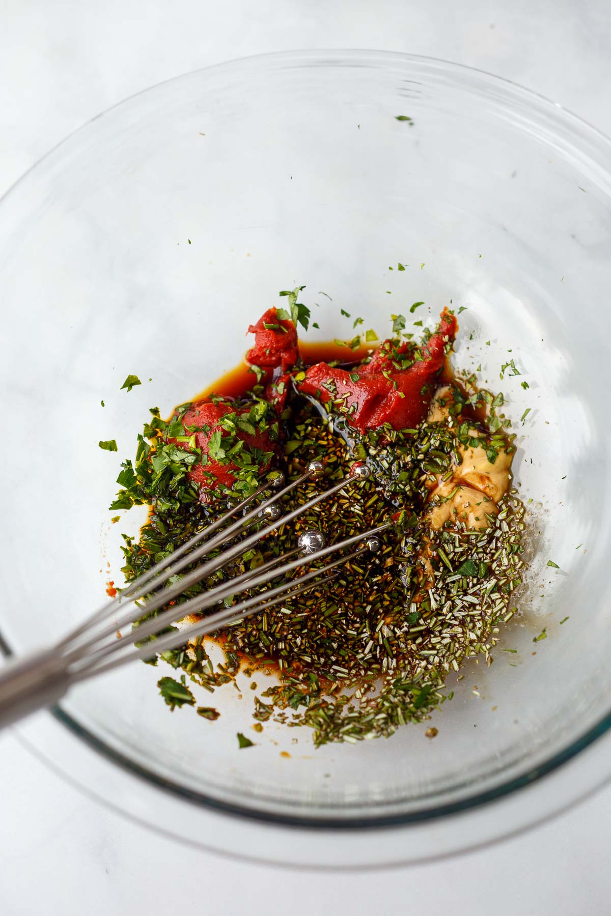 glass mixing bowl with flavor enhancers - tomato paste, soy sauce, balsamic, pepper, parsley, rosemary, salt, and mustard with whisk.