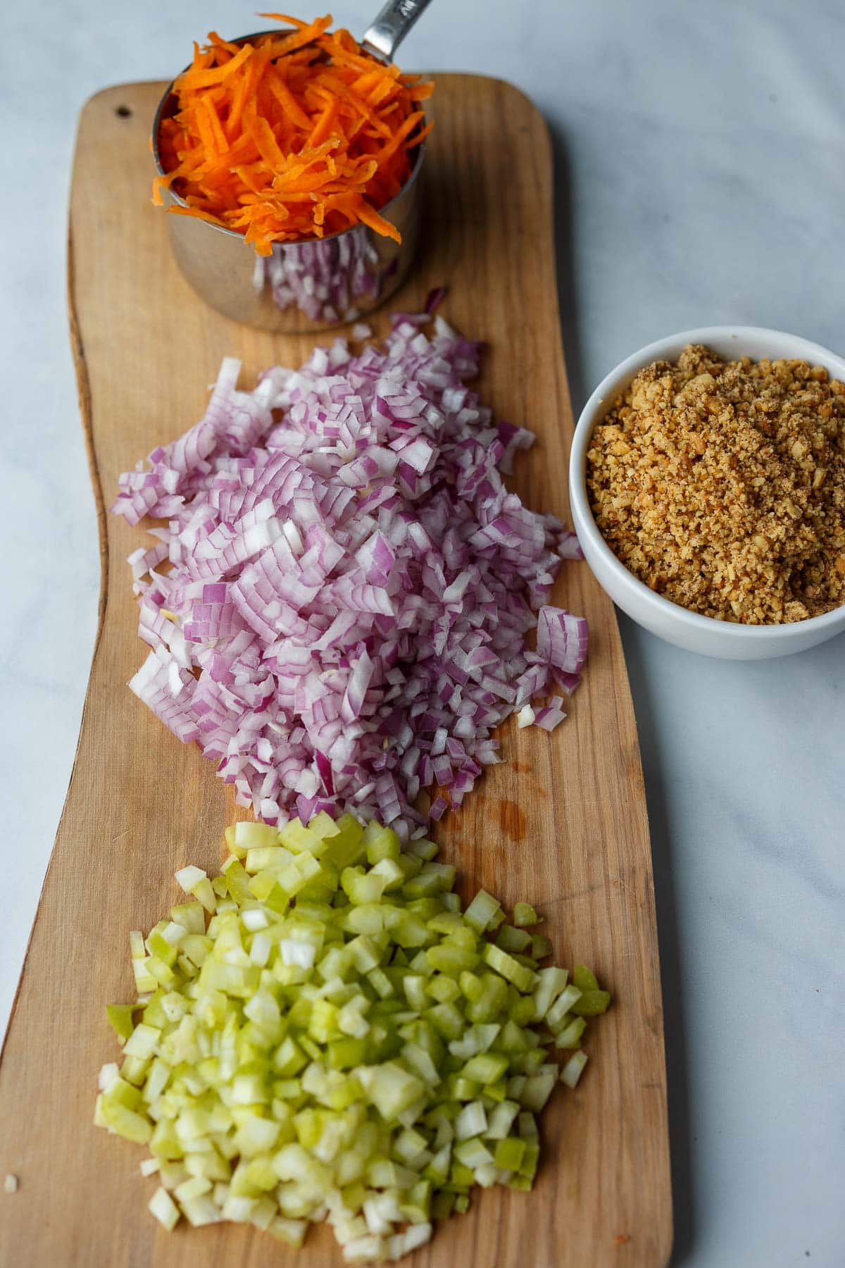 wood board with diced celery and onion and grated carrot, next to bowl of coarse walnuts.