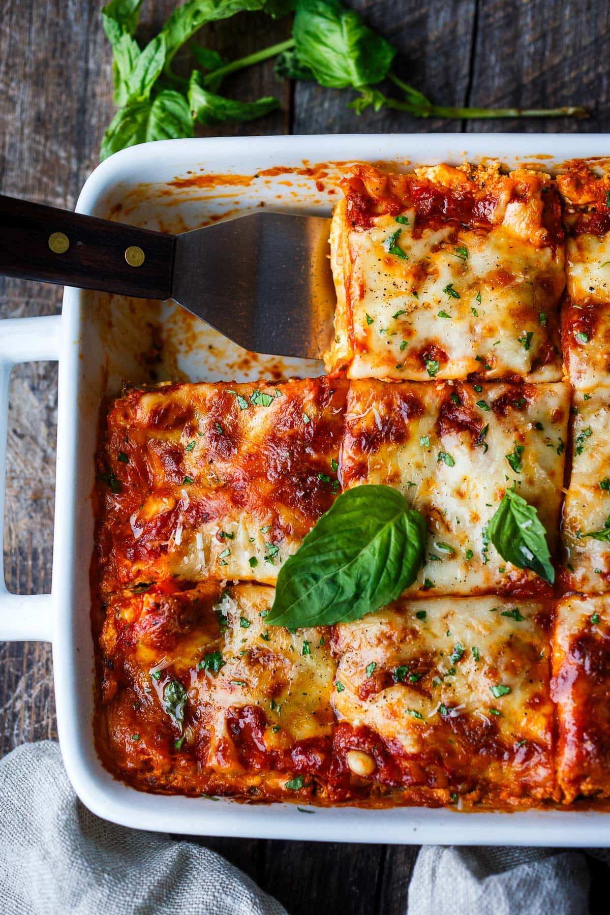 baking dish of golden brown cheesy vegetable lasagna garnished with fresh basil with spatula scooping out a square piece.