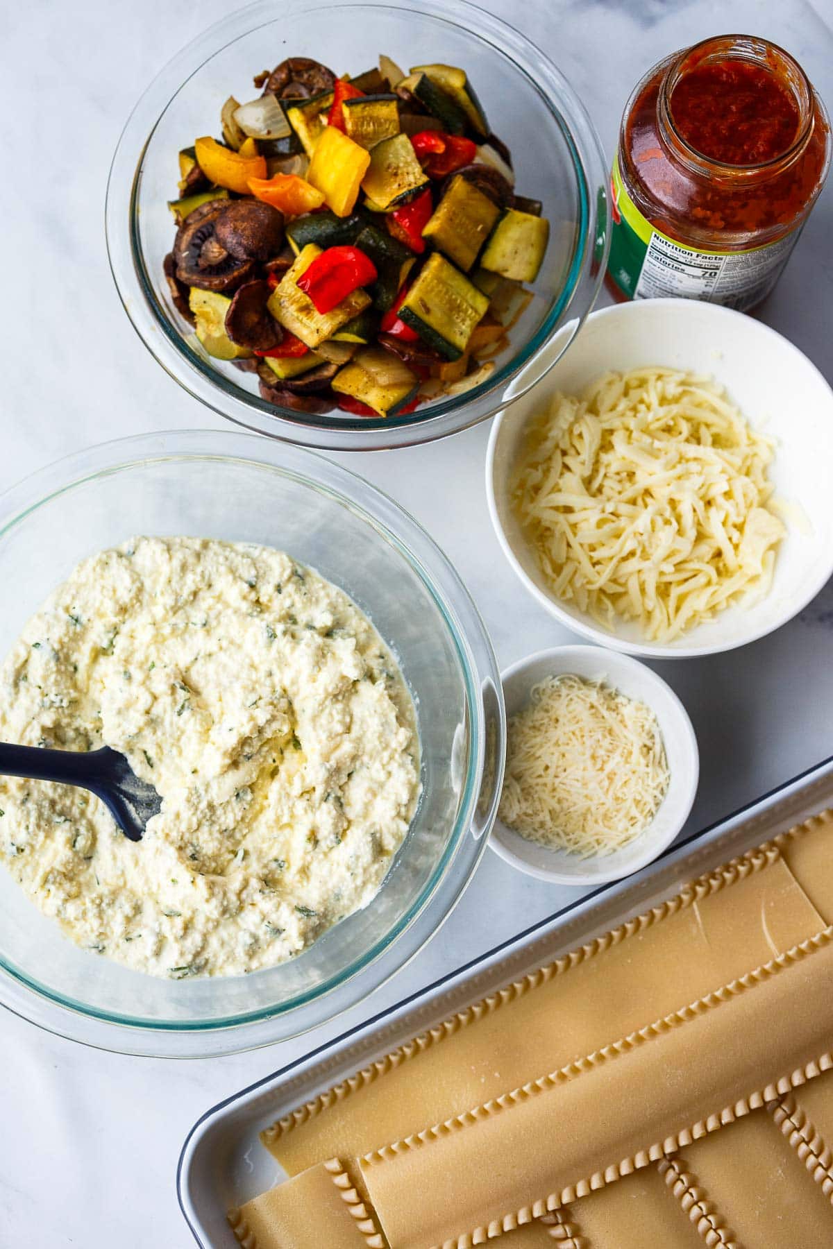 getting ready to assemble lasagna with jar of marinara, bowl of roasted veggies, and bowls of ricotta mixture, mozzarella, and parmesan, next to pan of lasagna noodles.