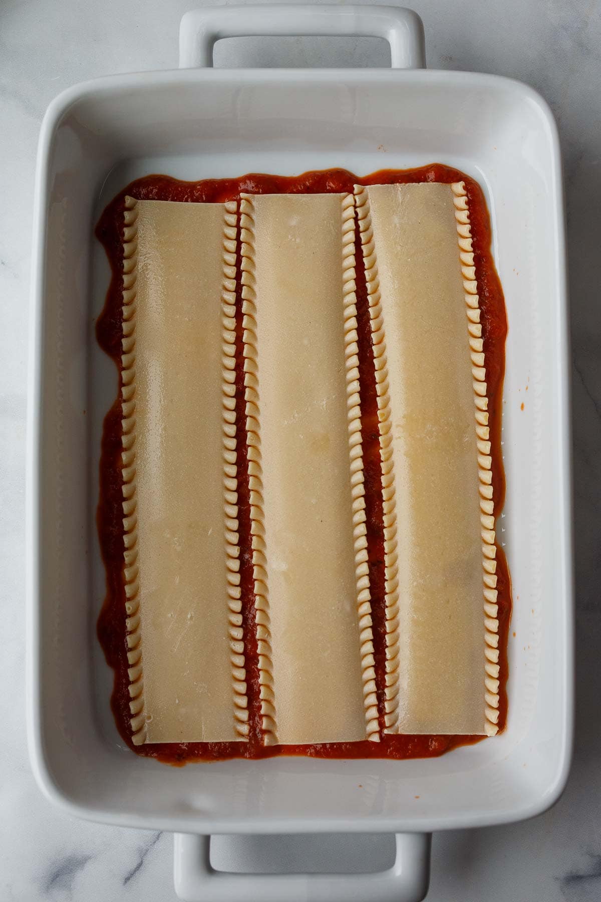 three lasagna noodles placed on top of marinara in casserole dish.