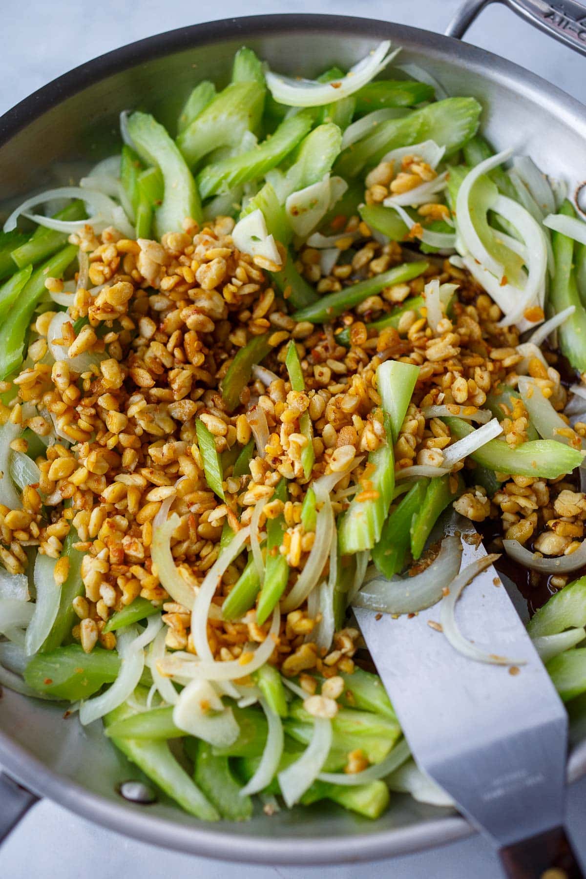 tempeh added to skillet with stir-fried celery and onion with metal spatula. 