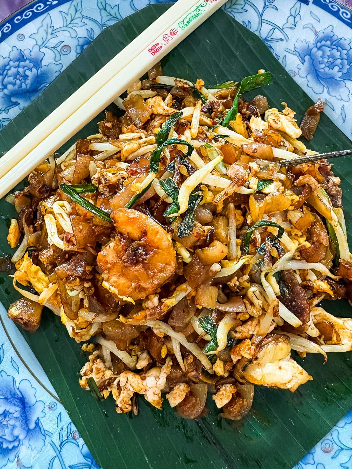 char kway teow on palm leaf-lined plate with chopsticks - made with prawns, bean sprouts, eggs, and chives.
