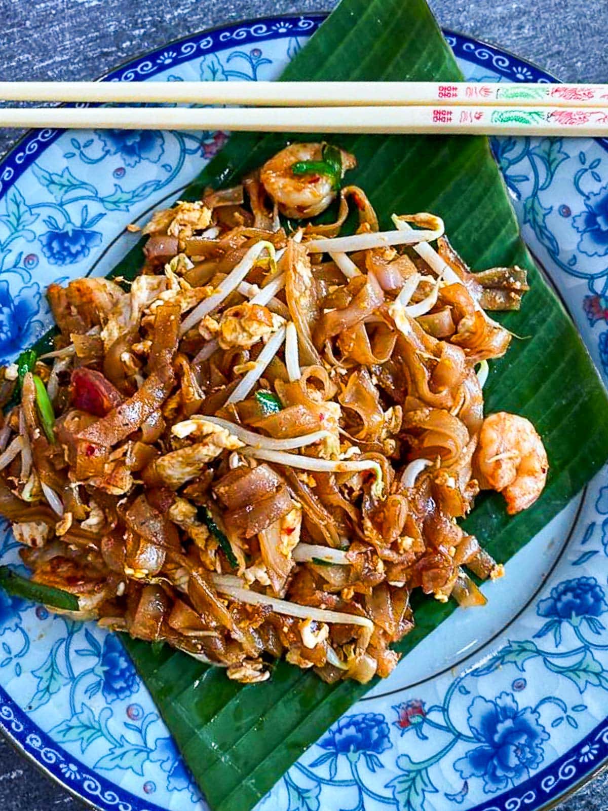 serving of char kway teow on palm leaf-lined blue plate with chopsticks.