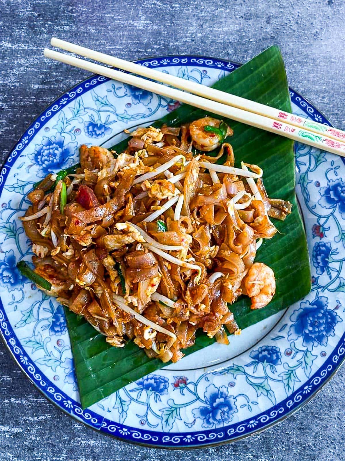 blue plate with palm leaf and chopsticks featuring char kway teow with bean sprouts and chives.