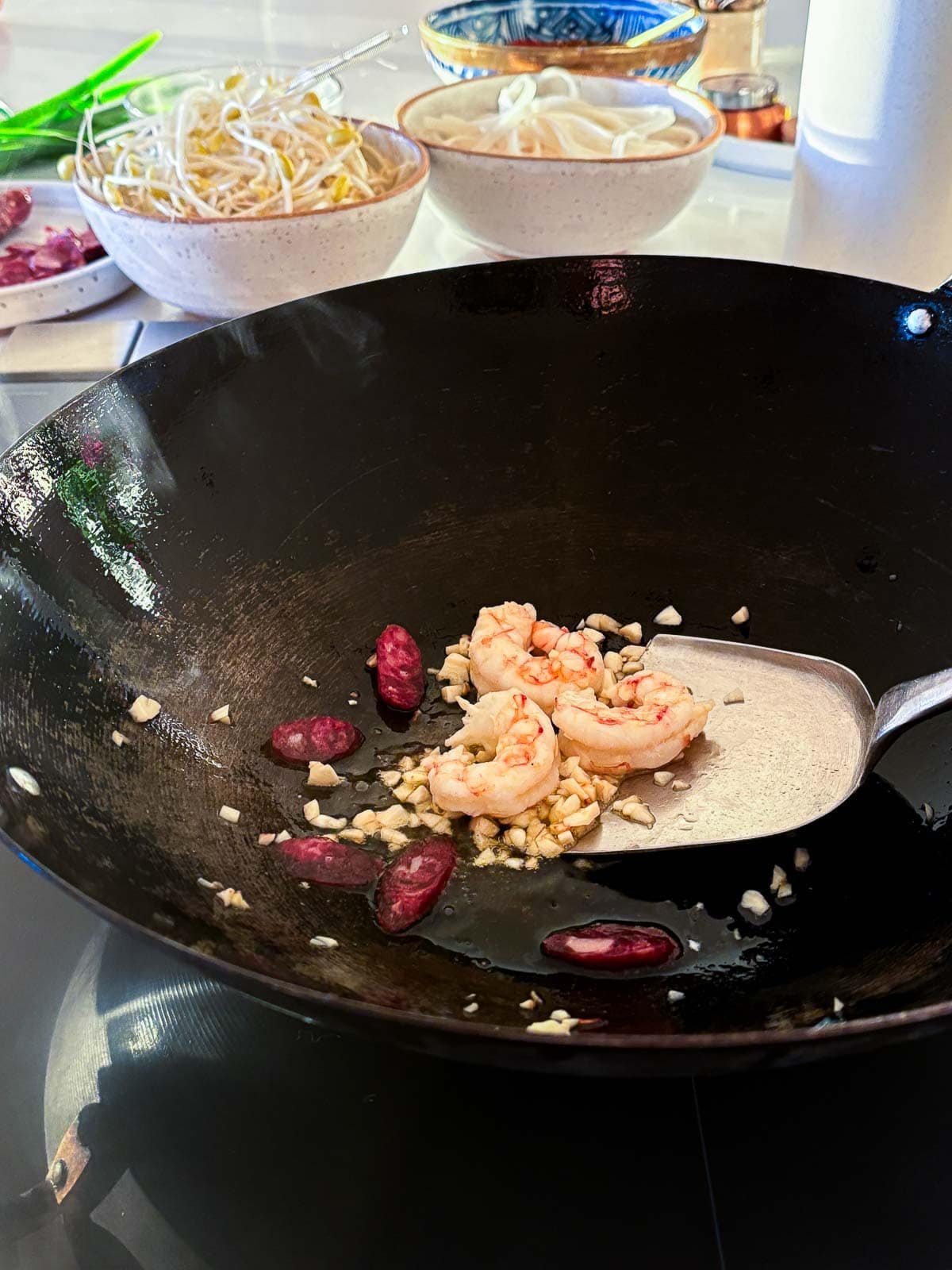 wok with metal spatula stirring prawns, garlic, and sausage in oil.