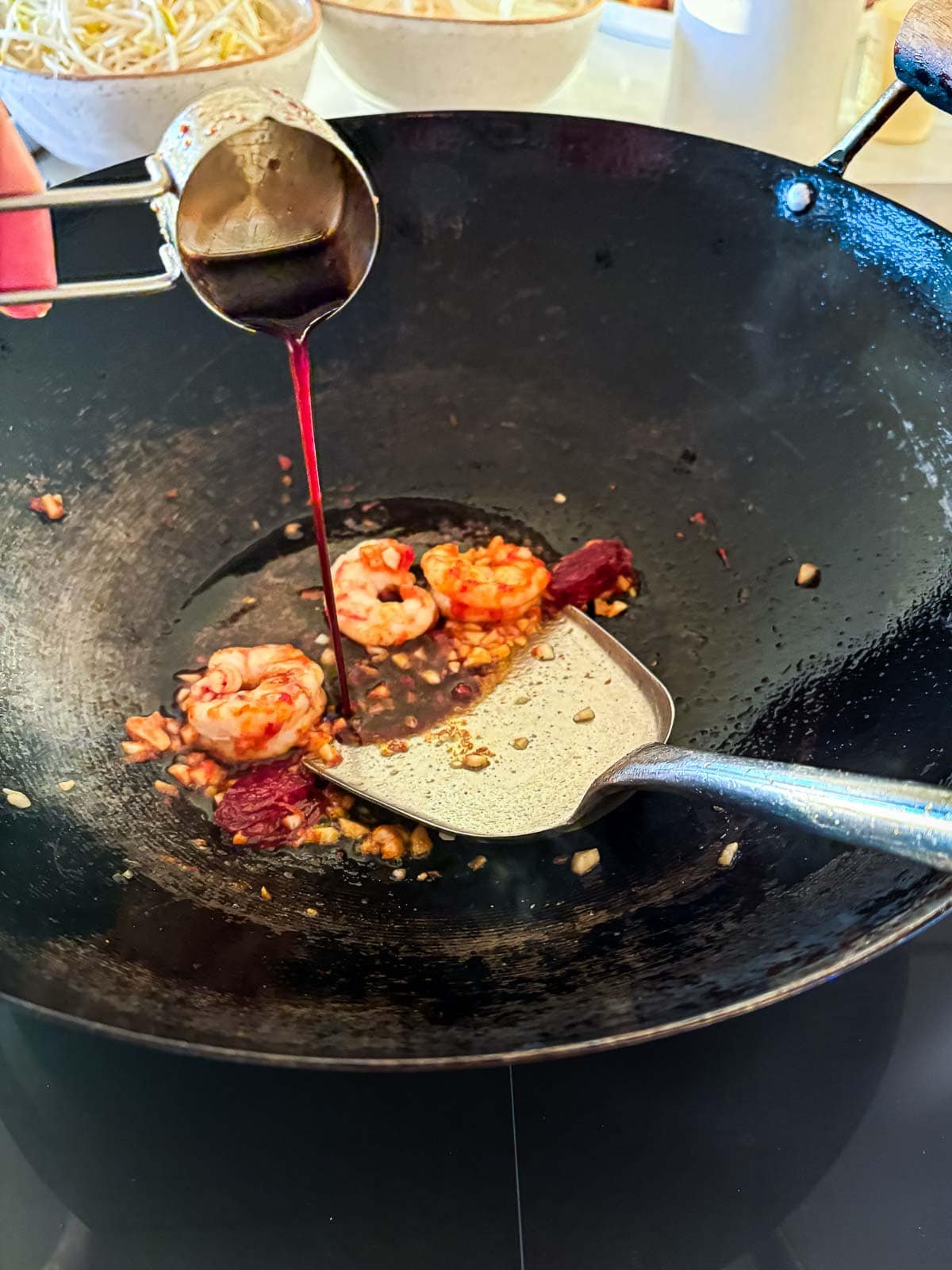 pouring stir fry sauce into wok with metal spatula, prawns, garlic, and Chinese sausage.
