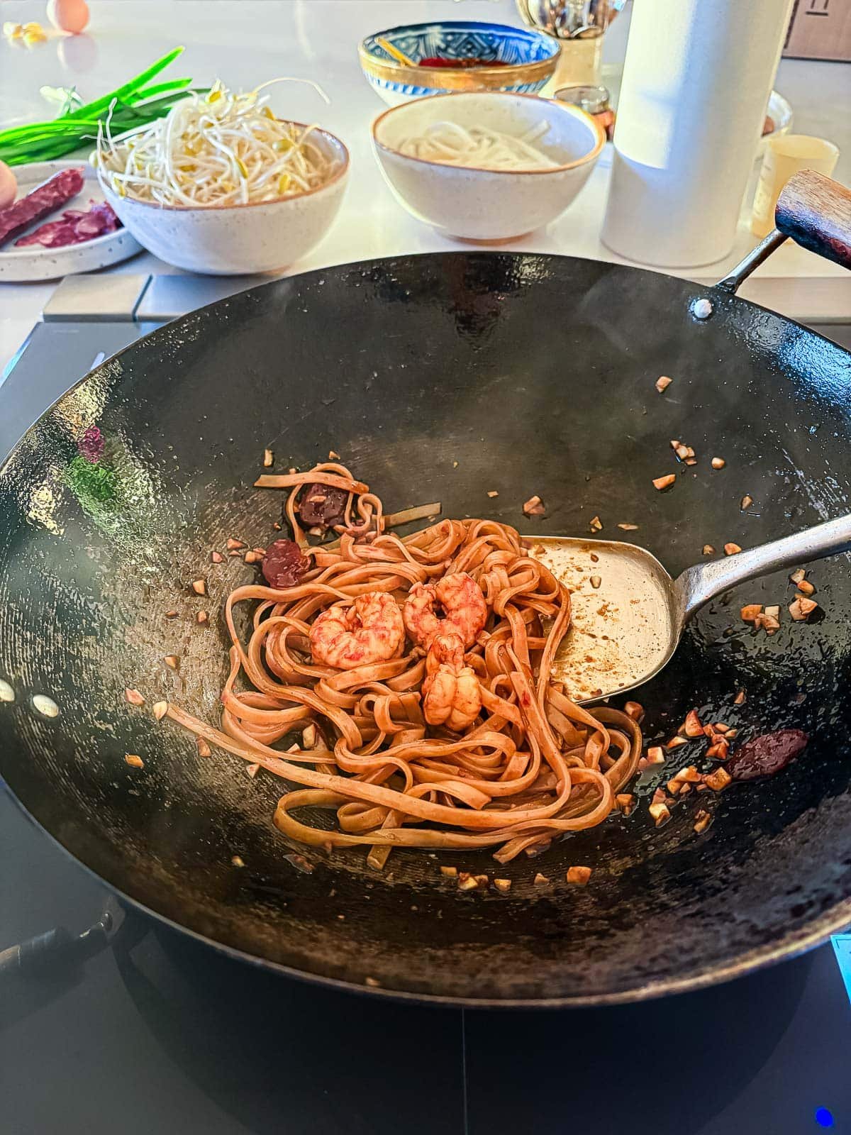 metal spatula stirring noodles and prawns in char kway teow stir fry sauce in wok.