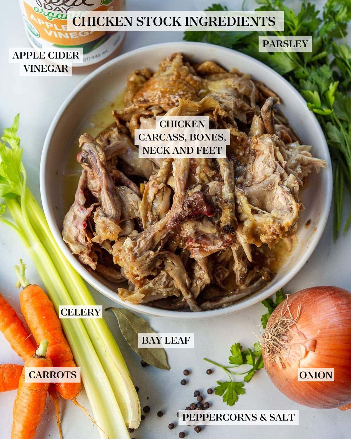ingredients for homemade chicken stock - bowl of roasted chicken carcass and bones, surrounded by parsley, onion, carrots and celery, bay leaf, peppercorns, and apple cider vinegar. 