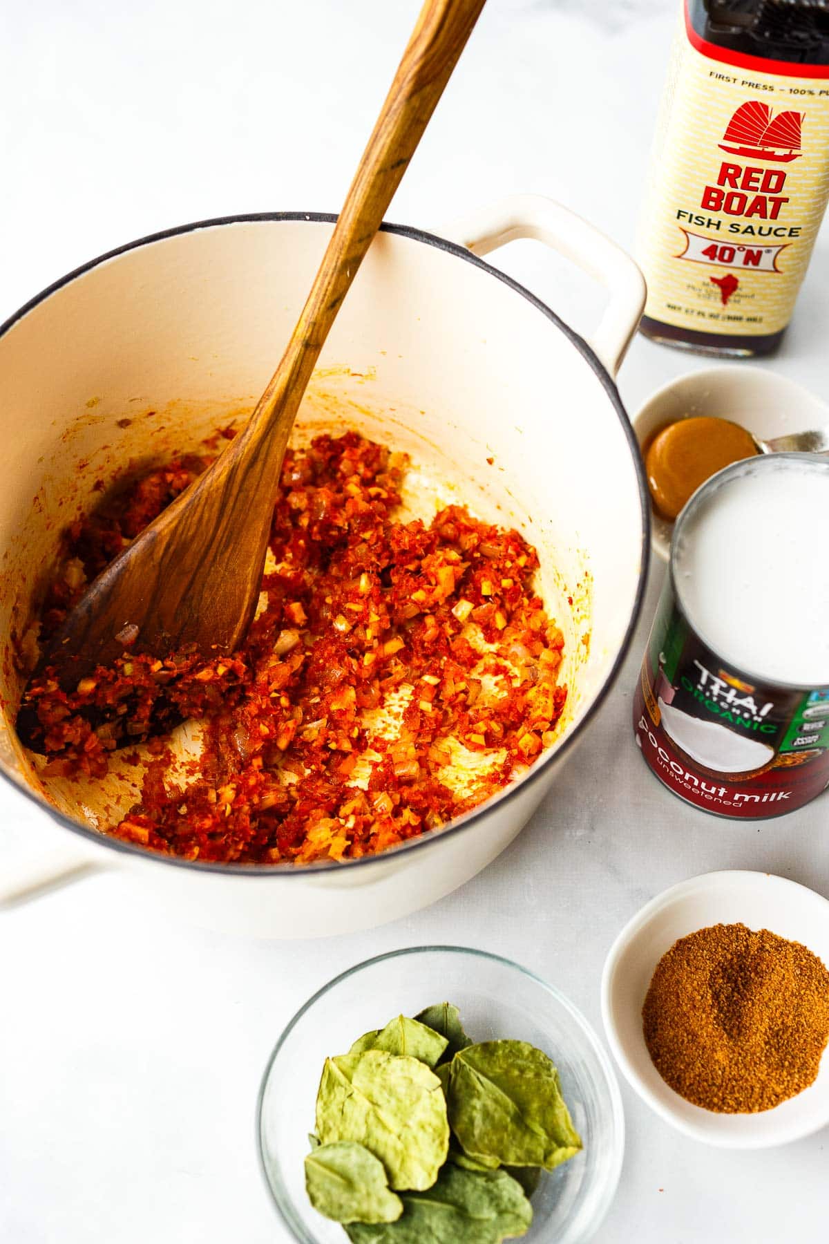 aromatics and panang curry paste stirring together with wooden spoon in dutch oven next to panang curry ingredients, kaffir lime leaves, coconut sugar, coconut milk, and fish sauce.
