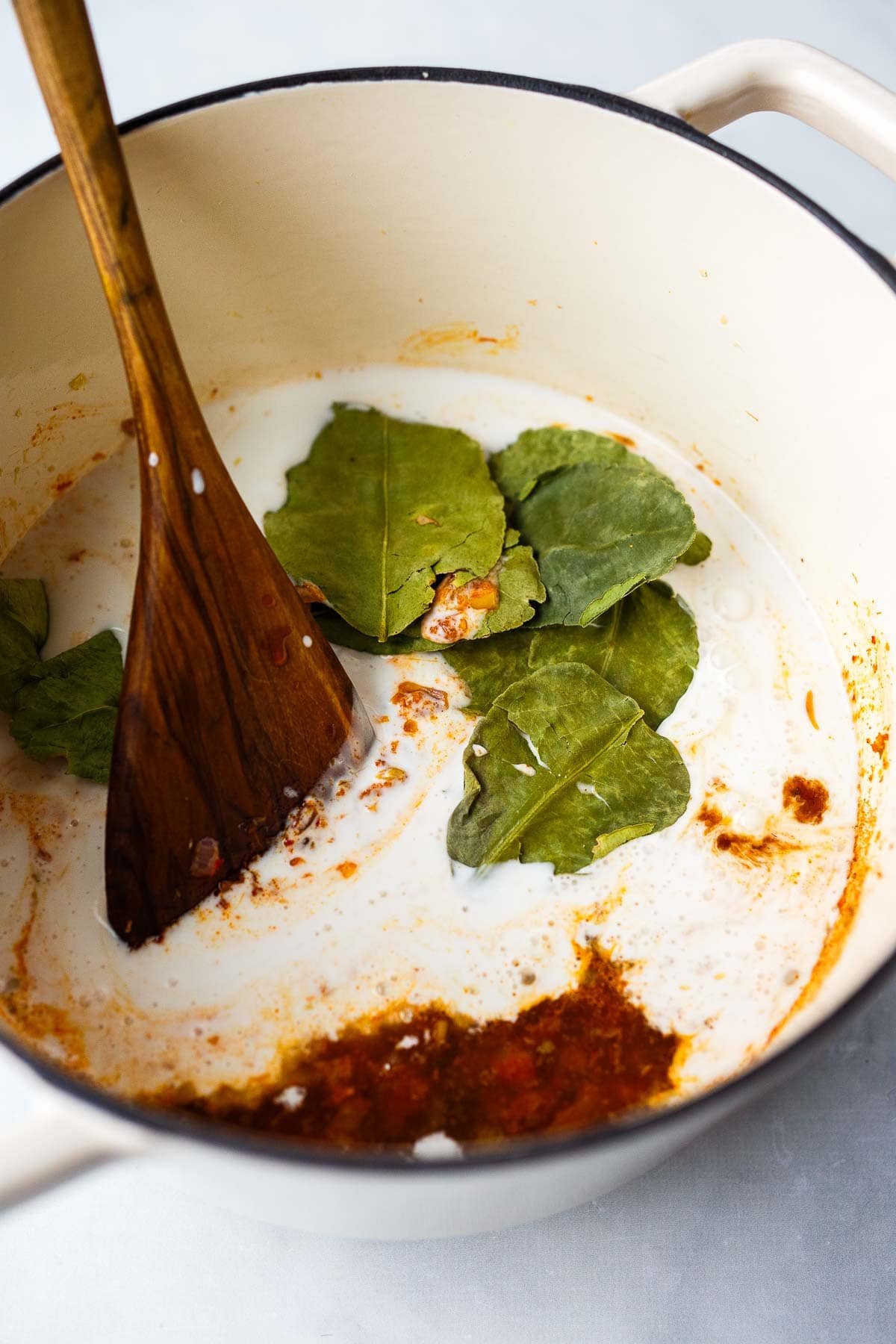 dutch oven with wood spoon stirring coconut milk into panang curry with kaffir lime leaves. 