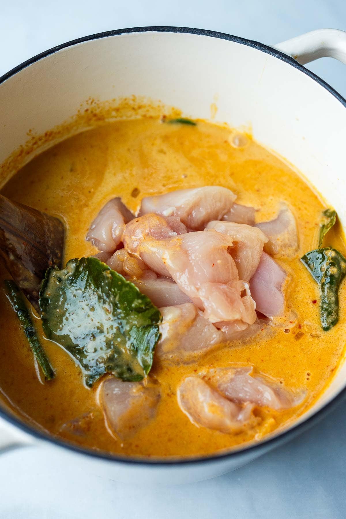 raw chicken cubes and thai basil leaves in creamy coconut panang curry in dutch oven. 