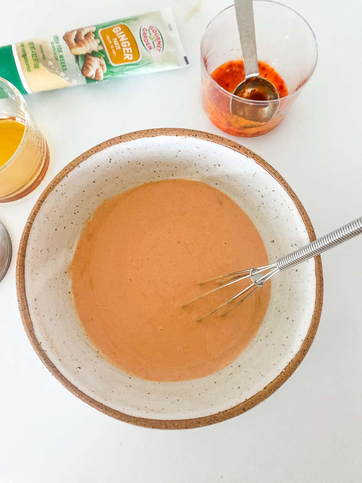 ceramic bowl with whisk stirring yum yum sauce next to jar of chili garlic sauce with measuring spoon. 