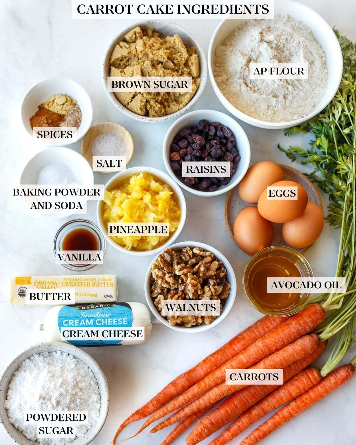 carrot cake ingredients arranged in bowls and labeled - ap flour, brown sugar, spices, salt, raisins, eggs, baking powder and baking soda, pineapple, avocado oil, vanilla, walnuts, butter, cream cheese, carrots, powdered sugar. 