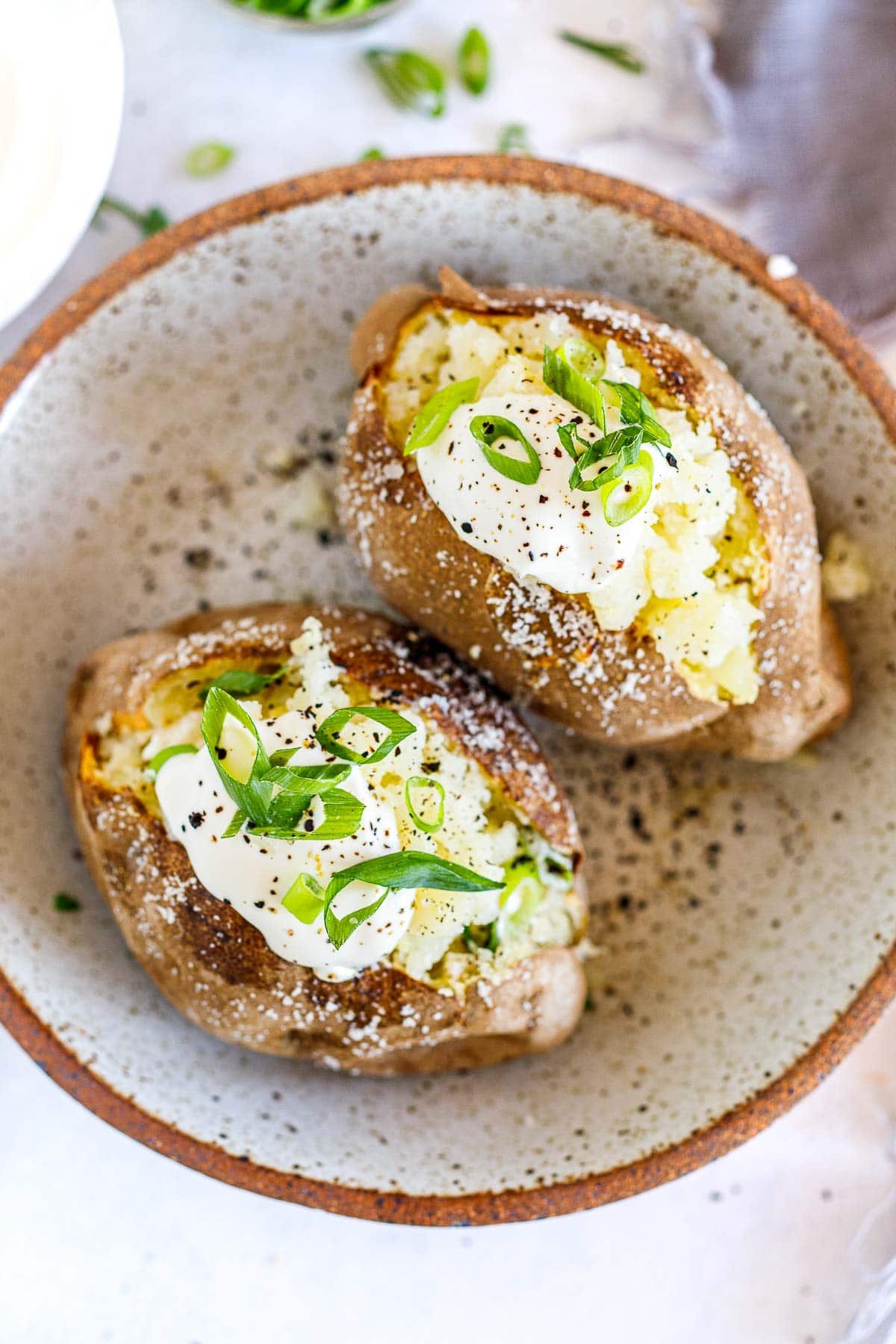 two baked potatoes topped with sour cream and green onions and red pepper flakes arranged side by side in a bowl. 
