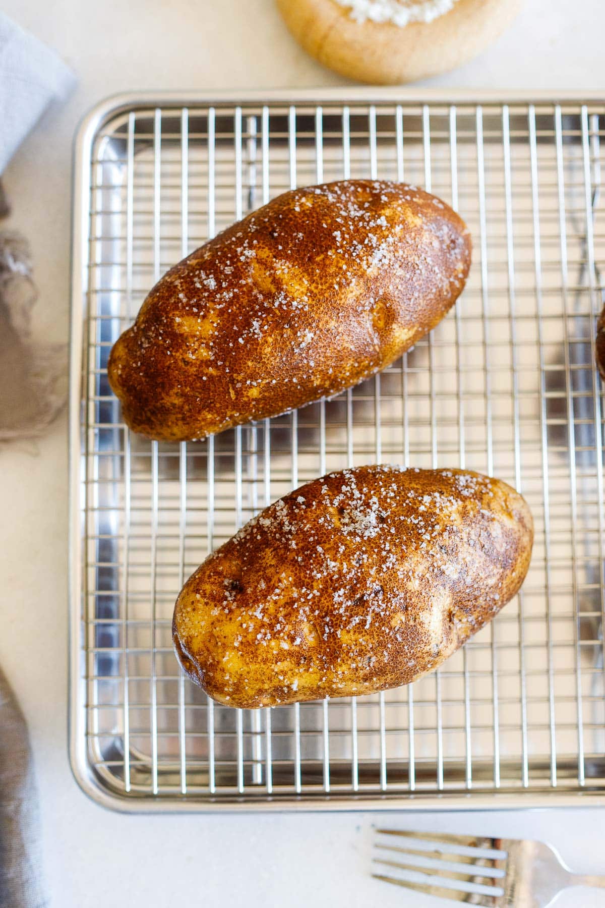 two russet potatoes coated in olive oil and seasoned with salt on a wire rack on a baking sheet. 