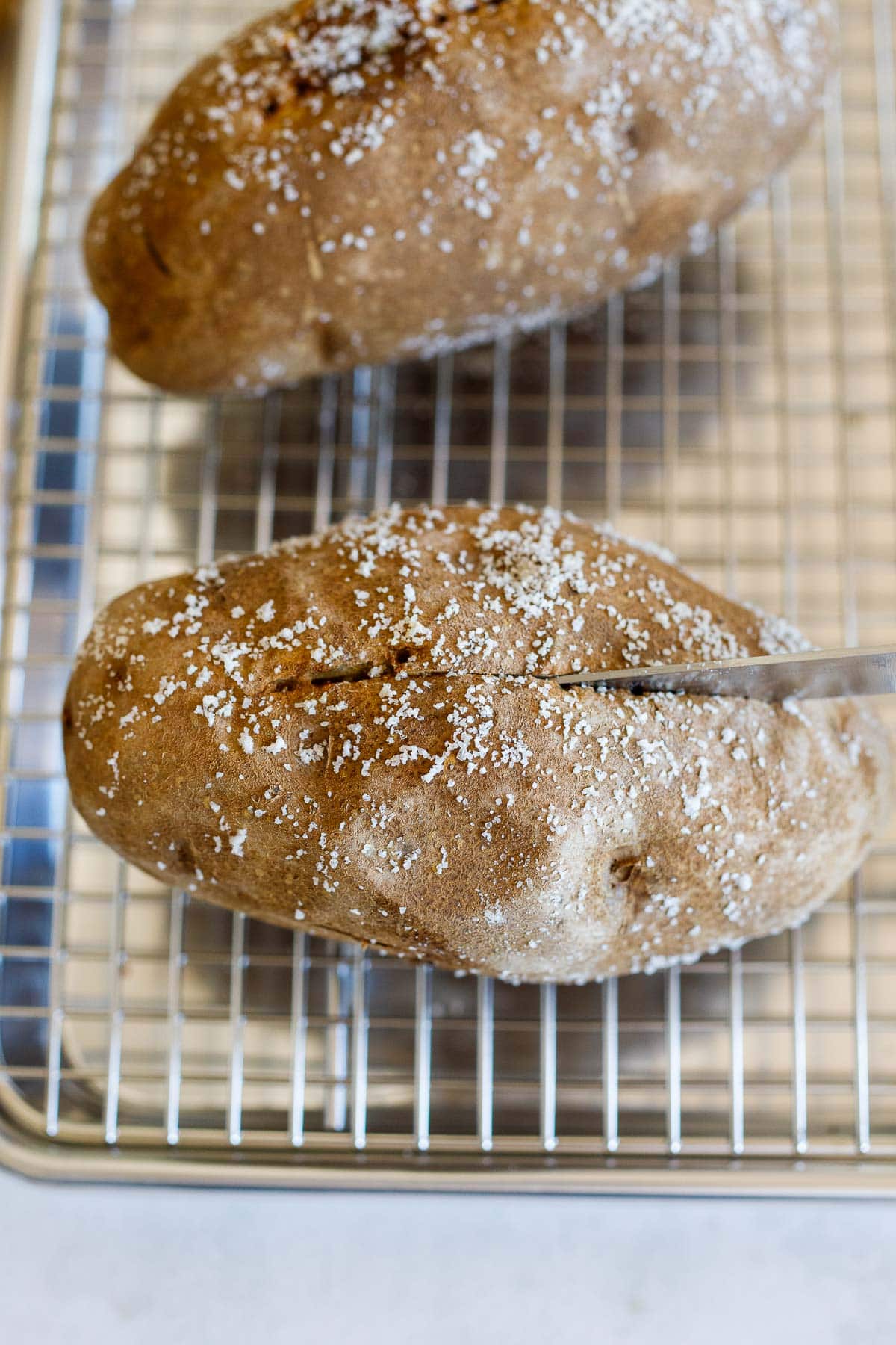 knife slicing the top of a baked potato while cooling on a wire rack.