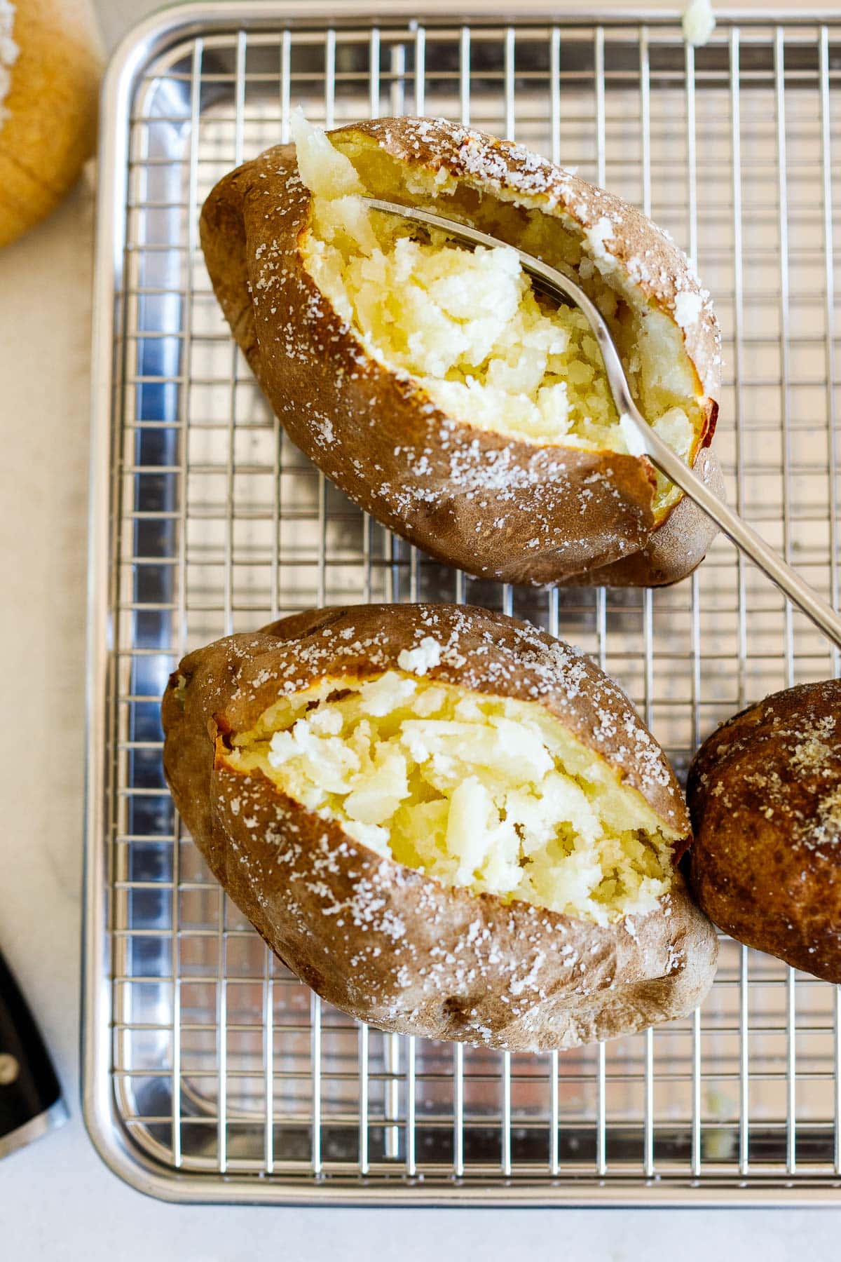 baked potato on wire rack with a fork laying down the center to reveal a fluffy interior, next to another baked potato that has been cut open.