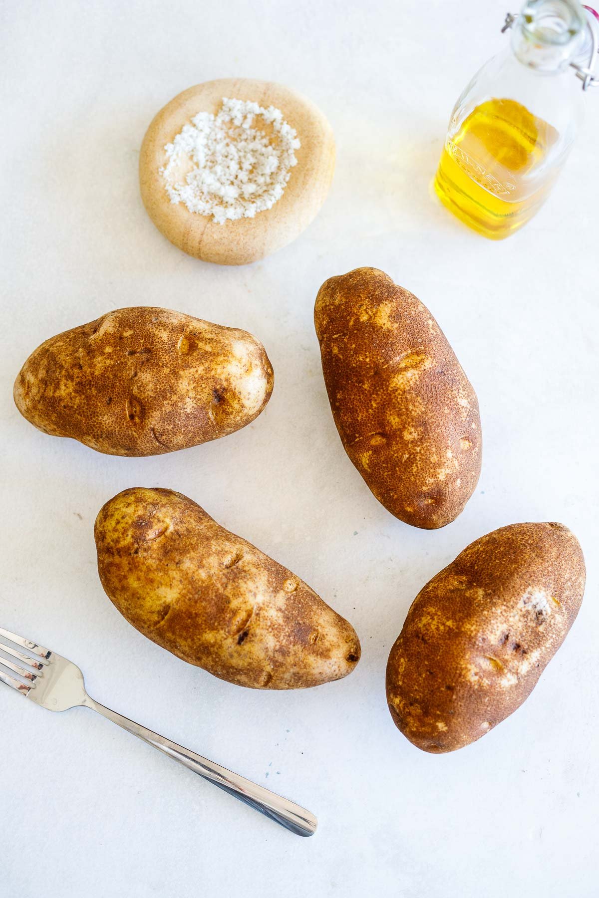 ingredients for baked potato - four russet potatoes surrounded by a fork, salt, and olive oil. 