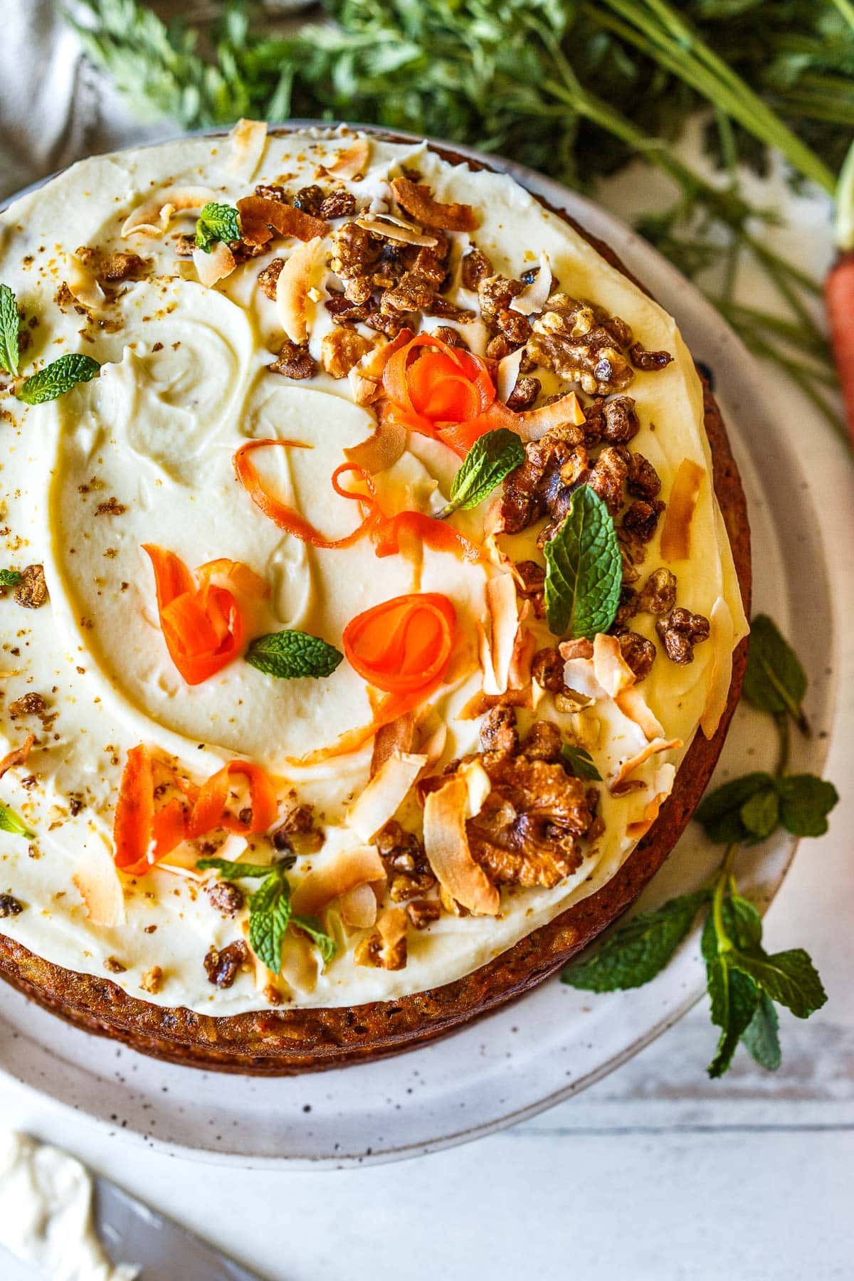 frosted carrot cake on serving plate garnished with carrot ribbons, mint, toasted walnuts, and toasted coconut. 