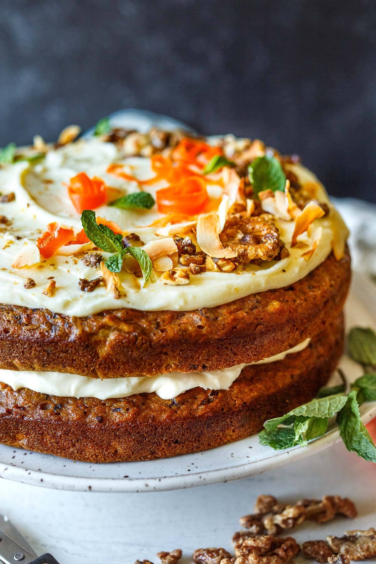 carrot cake with two round layers and cream cheese frosting in between, with the top layer garnished with mint leaves, toasted coconut and walnuts, and carrot ribbons.