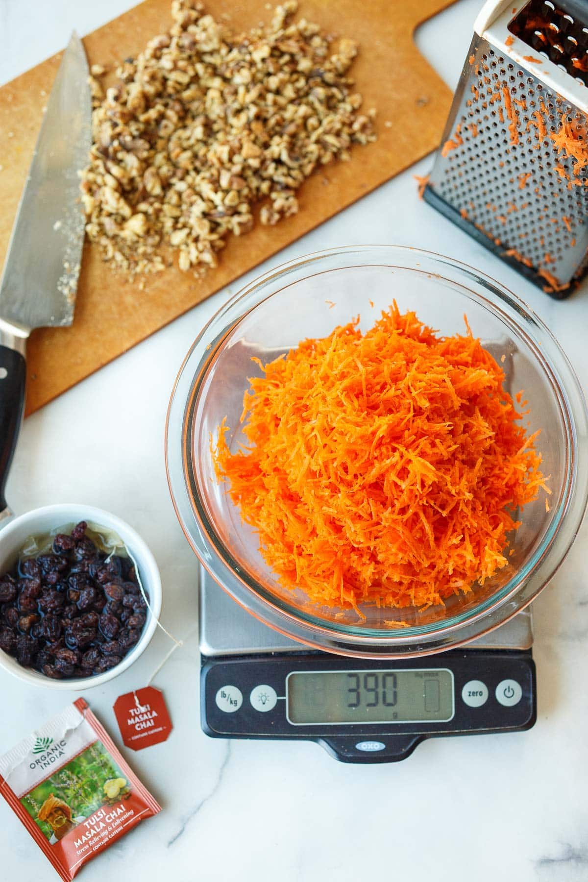 glass bowl of grated carrots on kitchen scale next to small bowl of raisins soaking in water with chai tea bag, and a cutting board with walnuts in the background. 