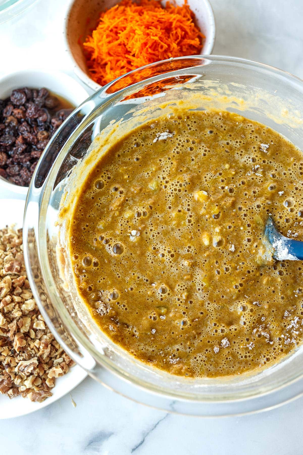 glass mixing bowl with wet ingredients surrounded by bowls of carrots, raisins, and walnuts.