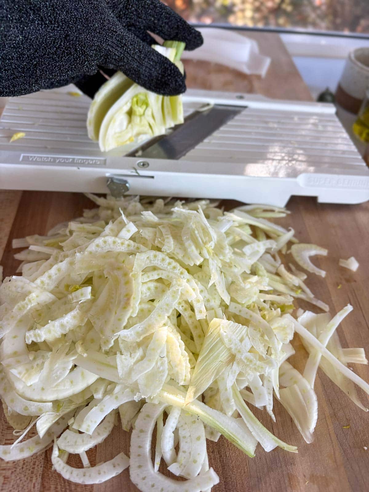 gloved hand running fennel down mandolin behind pile of sliced fennel on wood board. 