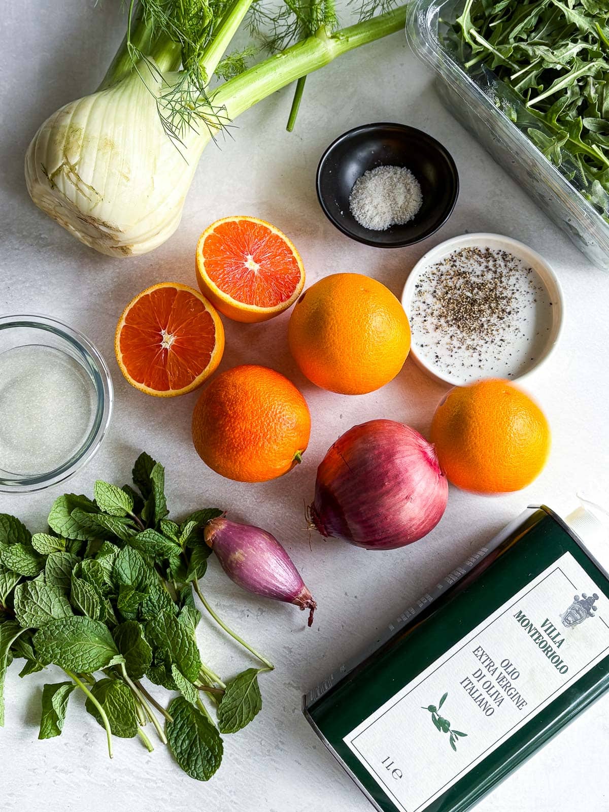 ingredients laid out for fennel orange salad - fennel, arugula, salt, pepper, oranges, shallot, red onion, olive oil, mint.